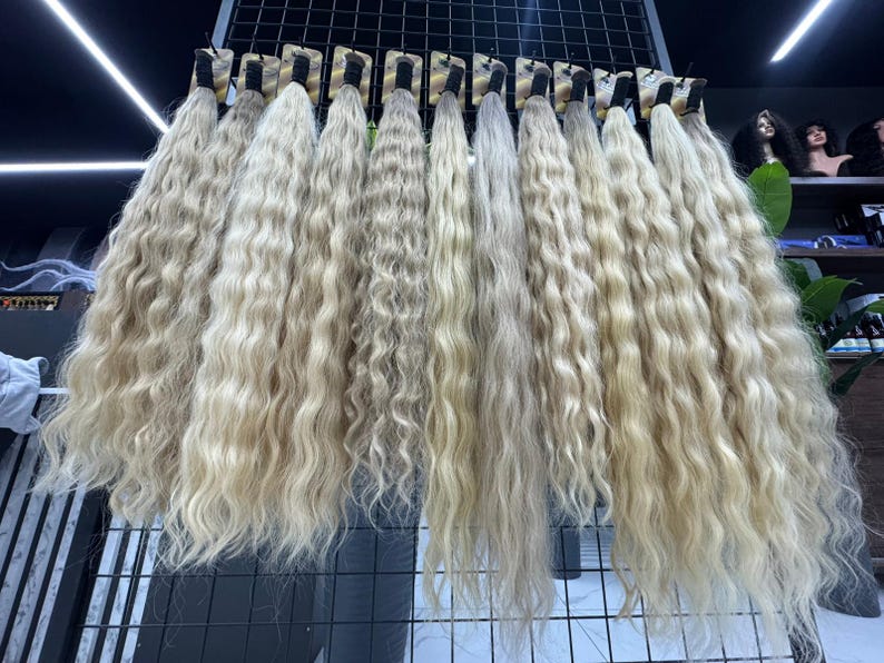 以下が含まれることがあります： ウェーブのかかったブロンドのヘアエクステンションの展示。暗いベースの各束は、金属製のラックに吊り下げられています。さまざまなブロンドの色合いと髪のウェーブのかかった質感がはっきりと見えます。