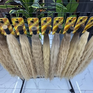 以下が含まれることがあります： ウェーブのかかったブロンドのヘアエクステンションのディスプレイ。各束は黒いバンドで固定されています。髪の色合いは明るいブロンドからミディアムブロンドまであり、黒い金属製のラックに配置されています。