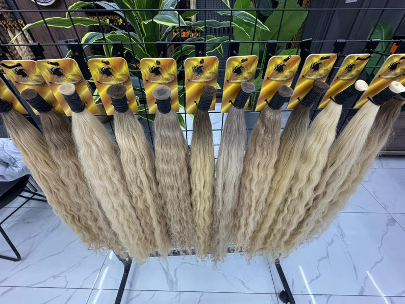 以下が含まれることがあります： ウェーブのかかったブロンドのヘアエクステンションのディスプレイ。各束は黒いバンドで固定されています。髪の色合いは明るいブロンドからミディアムブロンドまであり、黒い金属製のラックに配置されています。