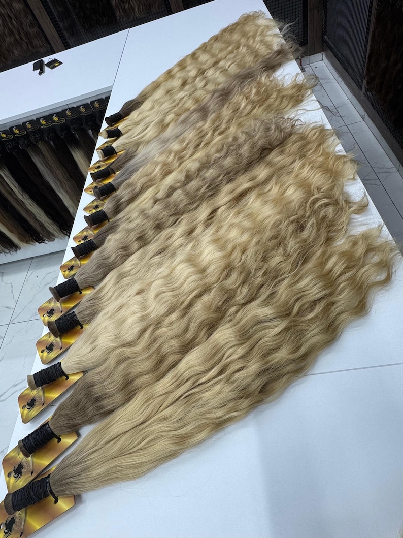 以下が含まれることがあります： 白い表面に並べられた、波状の明るいブロンドのヘアエクステンションの束がいくつかあります。髪は、根元にさまざまなブロンドと茶色の色合いがあり、自然な外観をしています。各束は黒い糸と金色のラベルで固定されています。