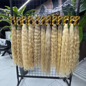 以下が含まれることがあります： 黒い金属製のラックに吊るされた、ブロンドのウェーブのかかったヘアエクステンションのディスプレイ。ヘアバンドルは、明るい色から中間の色まで、さまざまなブロンドの色合いがあります。各バンドルは、小さな金色のカードに添付されています。