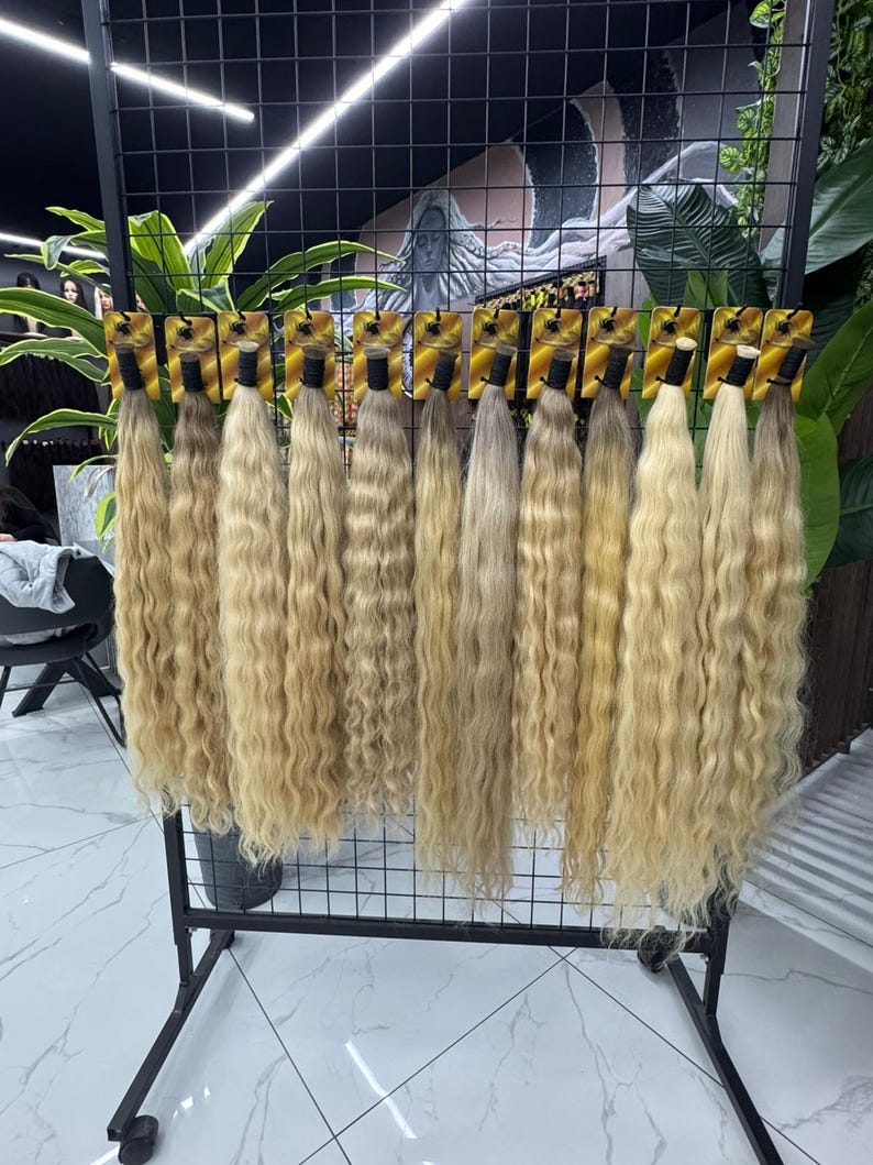 以下が含まれることがあります： 黒い金属製のラックに吊るされた、ブロンドのウェーブのかかったヘアエクステンションのディスプレイ。ヘアバンドルは、明るい色から中間の色まで、さまざまなブロンドの色合いがあります。各バンドルは、小さな金色のカードに添付されています。