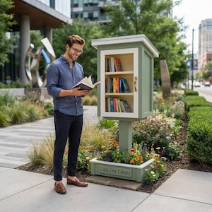 Peut inclure: Une boîte de partage de livres Little Free Library vert sauge avec une porte vitrée, deux étagères intérieures remplies de livres colorés et une base jardinière fleurie, située sur un trottoir en béton dans un cadre urbain.