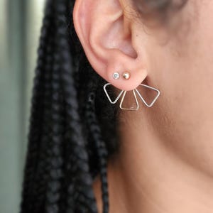 Peut inclure: Gros plan sur une oreille ornée de trois boucles d'oreilles. Les deux supérieures sont de petits clous, l'un avec une pierre transparente et l'autre une boule dorée. La troisième est un design géométrique argenté et doré.