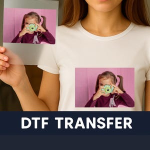 May include: White t-shirt featuring a printed image of a girl with a green camera. A clear transfer sheet with the same image is held in front of the shirt. The words "DTF TRANSFER" are printed in white on a dark blue background.