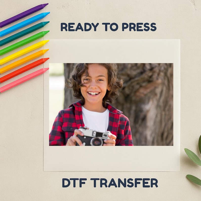 May include: A cream-coloured DTF transfer design with the text "READY TO PRESS" and "DTF TRANSFER". The image features a smiling person holding a vintage camera, wearing a red and black plaid shirt. Colourful crayons are in the upper left corner.