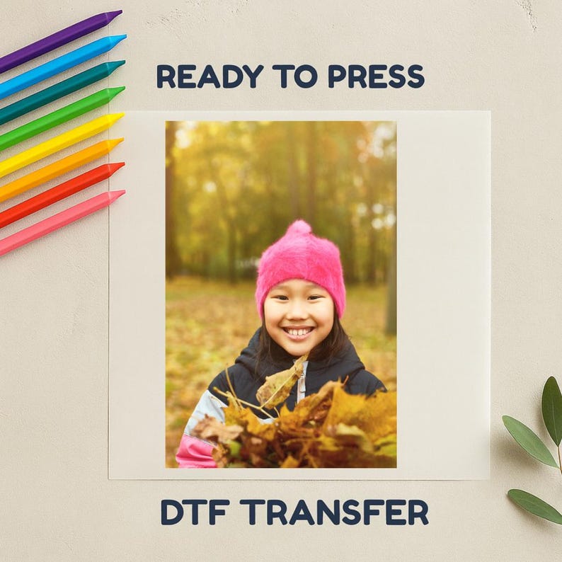 May include: A girl wearing a pink hat and holding autumn leaves, with the text "READY TO PRESS" and "DTF TRANSFER". Colour pencils are arranged along the top left.
