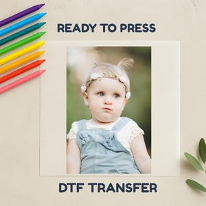 May include: A baby wearing a floral headband and denim dungarees. The image is labeled "READY TO PRESS" and "DTF TRANSFER". Crayons in various colours are arranged along the left side of the image.