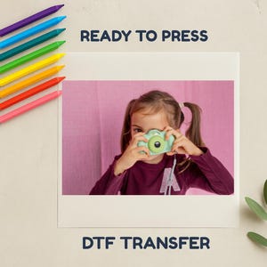 May include: A child holding a mint green toy camera, framed within a white border. The image is set against a pink backdrop, with the text "READY TO PRESS" and "DTF TRANSFER" above and below. Colourful crayons are visible in the upper left corner.