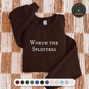May include: A dark brown crewneck sweatshirt featuring the white text Worth the Splinters printed on the chest. The garment has long sleeves and a ribbed collar. A small circular logo with a flame icon and the text Fourfiftyone is visible.