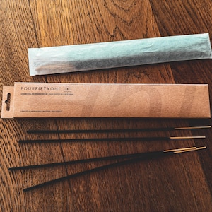 May include: Incense sticks with gold tips and a cardboard box that reads "FOURFIFTYONE CHARCOAL INCENSE STICKS | HAND DIPPED IN CALIFORNIA". The incense is black, and the box is brown. The packaging is on a wooden surface.