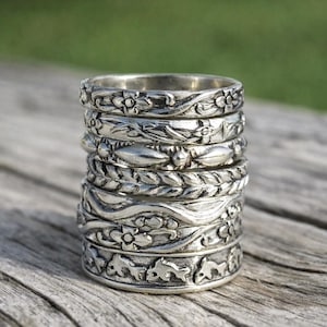 May include: A stack of silver rings with intricate floral and leaf patterns. The rings are stacked on a wooden surface, with a blurred green background. The rings have detailed designs, showcasing a variety of botanical motifs.