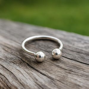 Puede incluir: Un anillo plateado con dos extremos esféricos. El anillo está hecho de un metal brillante y reflectante y descansa sobre una superficie de madera envejecida. El fondo es verde borroso.