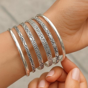 May include: A selection of silver bangles, some with detailed floral and scroll patterns, and others with a plain, polished finish. The bangles are stacked on a wrist, displaying the range of designs and textures.