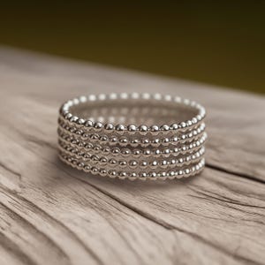 May include: A stack of six silver beaded bracelets. Each bracelet is composed of small, round beads. The bracelets are stacked on top of each other, creating a layered effect. The bracelets are on a wooden surface.