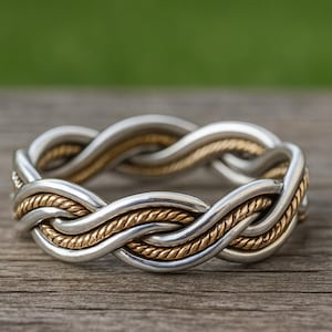 以下が含まれることがあります： シルバーとゴールドの編み込みリング。リングは、銀色のバンドと金色のロープのようなストランドが絡み合った織り模様が特徴です。リングは、ぼやけた緑と木の背景に配置されています。