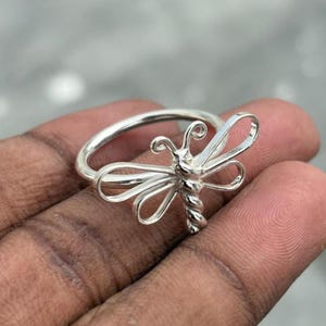 May include: A silver ring with a dragonfly motif. The dragonfly has detailed wings and a textured body. The ring is made of a shiny, metallic material. This is a piece of jewellery.