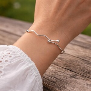Puede incluir: Un brazalete plateado con un diseño ondulado y dos pequeños detalles esféricos. El brazalete se lleva en la muñeca, con una camisa blanca de fondo. Una pieza de joyería.