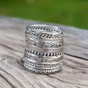 May include: A stack of silver rings with various textures and designs. The rings include braided, leaf, and patterned bands. The rings are displayed on a wooden surface, with a blurred green background.