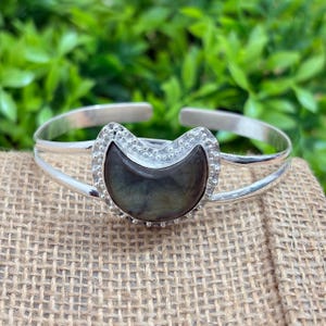 Könnte beinhalten: Ein silberfarbenes Armband mit einem zentralen, halbmondförmigen Stein in dunklen, irisierenden Grau- und Brauntönen. Der Stein ist von einem dekorativen, perlenartigen Metallrand umgeben, und das Band besteht aus zwei parallelen silberfarbenen Drähten.
