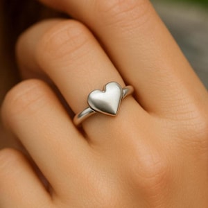 Könnte beinhalten: Ein silberner Ring mit herzförmigem Design. Das Herz ist glatt und massiv, mit abgerundeter Form. Der Ring ist schmal und schlicht, passend zum Herz. Der Ring wird am Finger getragen.