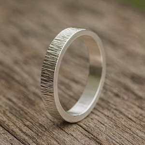 May include: A silver ring with a textured, bark-like surface. The ring has a simple band design and a polished interior. The ring is shown on a wooden surface.