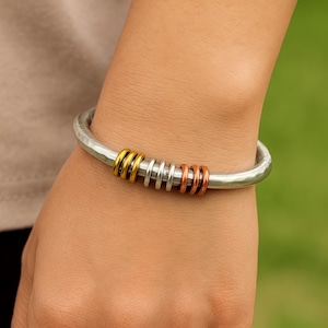 May include: A silver-toned bangle bracelet with a hammered texture. The bracelet features a series of colorful rings: gold, silver, and copper. The bracelet is worn on a wrist, showcasing the design and colors.