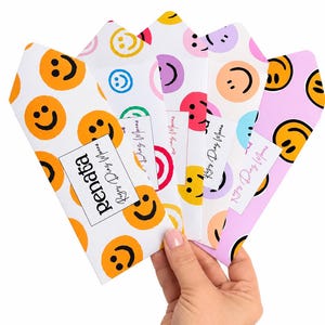 May include: A set of five paper envelopes featuring various smiley face patterns in orange, yellow, blue, pink, and purple. Each envelope displays the text Renata Reyes Diaz Morena on a white rectangular label attached to the front surface.