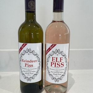 Op de afbeelding: Twee wijnflessen met donkere doppen en etiketten met de tekst "Reindeer Piss" en "Elf Piss". De etiketten hebben een decoratieve rand en de tekst "Bottled in the North Pole, Best Consumed When Already Pissed."