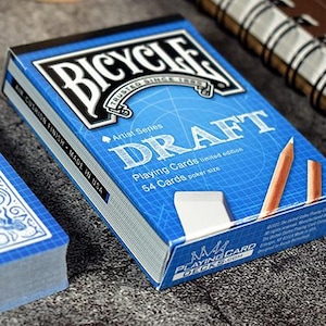 May include: A deck of Bicycle Draft playing cards, featuring a blue and white design. The box displays the word "DRAFT" and includes pencils and an eraser. The deck is a limited edition, with 54 cards in poker size.