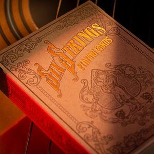 May include: A deck of "Big Rings Playing Cards" in a decorative box, resting on a guitar. The box is tan with gold lettering and ornate designs. The edges of the box are red. The guitar is brown and yellow.