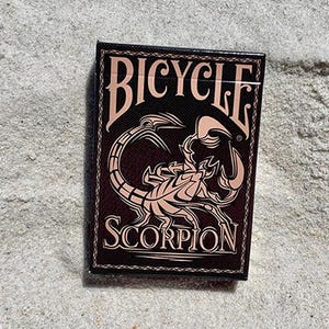 Puede incluir: Una baraja de cartas Bicycle Scorpion sobre arena. La caja de cartas es negra con una ilustraci&oacute;n de escorpi&oacute;n rosa y la palabra "SCORPION" debajo. La palabra "BICYCLE" est&aacute; arqueada sobre el escorpi&oacute;n.