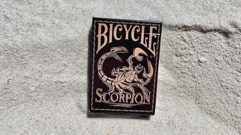Puede incluir: Una baraja de cartas Bicycle Scorpion sobre arena. La caja de cartas es negra con una ilustraci&oacute;n de escorpi&oacute;n rosa y la palabra "SCORPION" debajo. La palabra "BICYCLE" est&aacute; arqueada sobre el escorpi&oacute;n.