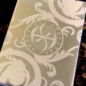 Puede incluir: Una caja de naipes plateada con un diseño floral blanco en relieve. El centro de la caja presenta un emblema circular con un diseño ornamentado. La caja tiene un brillo metálico.