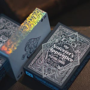 May include: A close-up shot of a deck of playing cards labelled "The Sorcerer's Apprentice." The cards are dark blue with white detailed designs. The Ace of Spades card has a holographic edge. The card box says "Playing Cards."