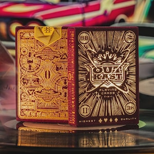 Puede incluir: Dos barajas de cartas en burdeos y dorado. Los dorsos de las cartas presentan diseños detallados con el texto "OUT KAST" y "PLAYING CARDS". Las caras de las cartas tienen un diseño dorado con temática alienígena.