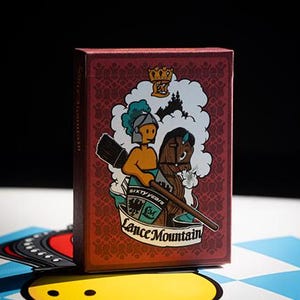 May include: A maroon playing card box with a decorative pattern and a colorful illustration. The illustration features a knight on a horse with the text "Sixty Years" and "Lance Mountain". The box is on a patterned surface.