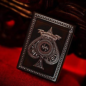 Puede incluir: Una baraja de cartas negra con un diseño de pica plateada. La pica es adornada con una corona y un pergamino. La baraja tiene un borde plateado y está sobre una superficie roja.