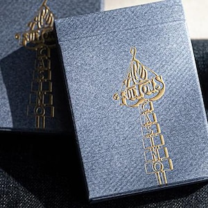 May include: Two gray playing card boxes with gold foil lettering and design. The boxes have a textured appearance. The gold design includes the words "ALLIONS" and other text. The boxes are on a dark gray surface.