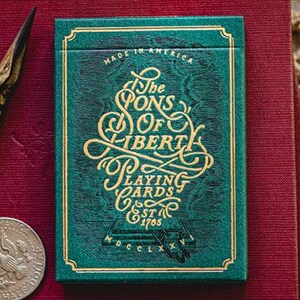 May include: A teal playing card box with gold lettering reading "The Sons of Liberty Playing Cards". The box is on a burgundy surface, with a silver ring, a vintage pen, and a silver coin. The text "Made in America" is at the top.