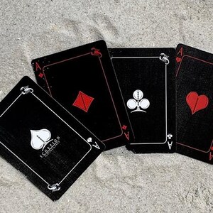 Puede incluir: Cuatro cartas de juego negras, cada una con el As de un palo diferente: picas, diamantes, tr&eacute;boles y corazones. Las cartas tienen detalles blancos y s&iacute;mbolos de palo rojos. Las cartas est&aacute;n sobre una superficie arenosa.