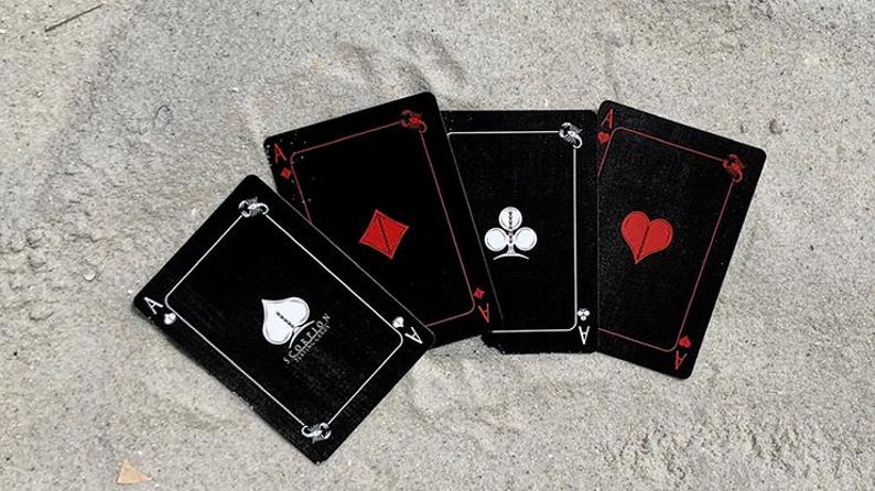 Puede incluir: Cuatro cartas de juego negras, cada una con el As de un palo diferente: picas, diamantes, tr&eacute;boles y corazones. Las cartas tienen detalles blancos y s&iacute;mbolos de palo rojos. Las cartas est&aacute;n sobre una superficie arenosa.