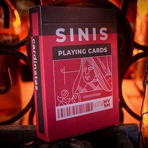 May include: A red and black deck of "SINIS PLAYING CARDS" with a white illustration of a queen. The card box has a barcode and the text "cardinates" on the side. The background is a blurred, warm-toned setting.