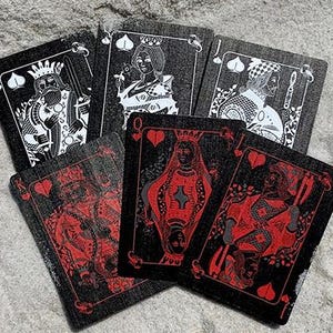 Puede incluir: Una baraja de cartas con fondo negro e ilustraciones rojas y blancas. Las cartas est&aacute;n abanicadas sobre una superficie texturizada de color gris claro. Los palos son visibles, incluyendo corazones y picas.