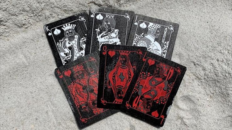 Puede incluir: Una baraja de cartas con fondo negro e ilustraciones rojas y blancas. Las cartas est&aacute;n abanicadas sobre una superficie texturizada de color gris claro. Los palos son visibles, incluyendo corazones y picas.