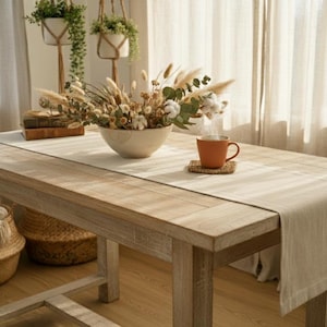 Op de afbeelding: Een rustieke houten tafel met een beige tafelloper, een keramische kom met gedroogde bloemen en een terracotta mok. Twee hangende plantenbakken met groen zijn zichtbaar op de achtergrond. Een gezellig, neutraal interieur.