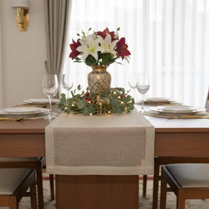 May include: A dining table set for a meal, featuring a light brown wooden table with a beige runner. The table is set with white plates, gold utensils, and wine glasses. A vase of red and white flowers sits in the center, with a small gold deer figurine.
