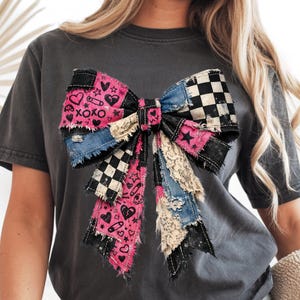 Op de afbeelding: Donkergrijs t-shirt met een groot strikdesign. De strik is een patchwork van denim, roze stof met hartjes en XOXO, zwart-wit geblokt en kant. De strik heeft een versleten, vintage uitstraling.