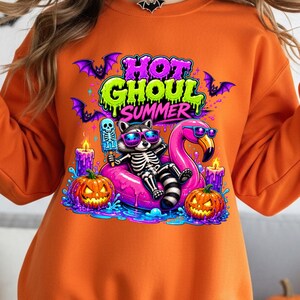 May include: Orange sweatshirt with a colorful graphic. The graphic features a skeleton raccoon wearing sunglasses, lounging on a pink flamingo float, and holding a skeleton popsicle. The text "HOT GHOUL SUMMER" is above the raccoon, with bats and jack-o'-lanterns.