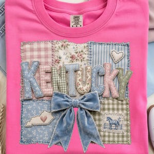 May include: Pink t-shirt with a patchwork design featuring the word "Kentucky" in various patterned fabrics. A blue velvet bow is centered below the text. The design includes a heart and a horse.